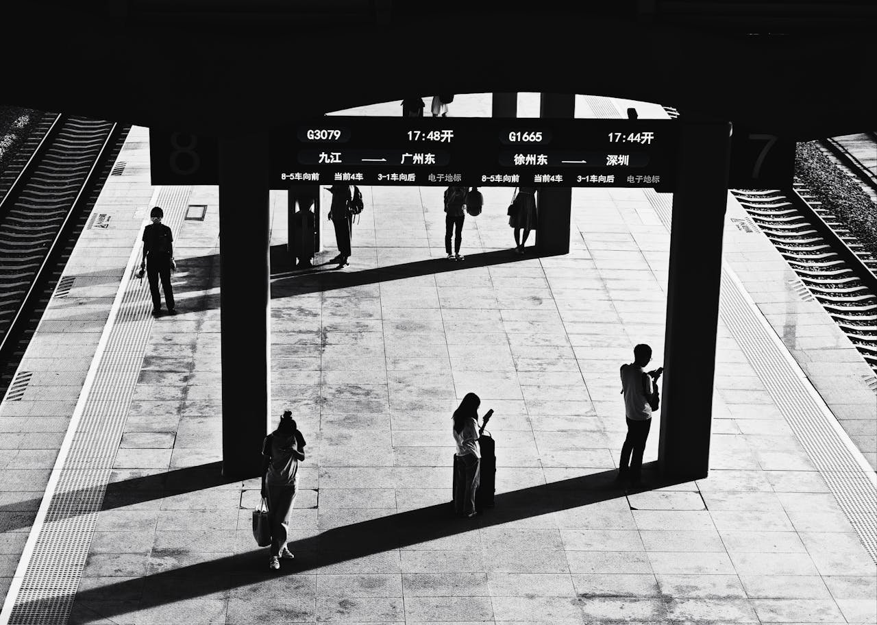 Monochrome image of a train station with people waiting, creating dramatic shadows.