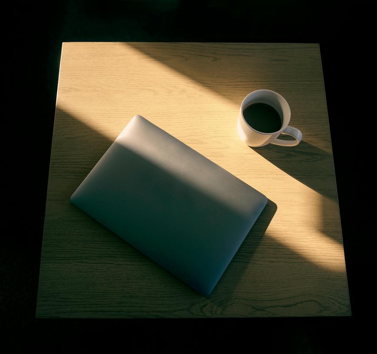 A minimalist setup featuring a laptop and a cup of coffee on a wooden table with light and shadow play.